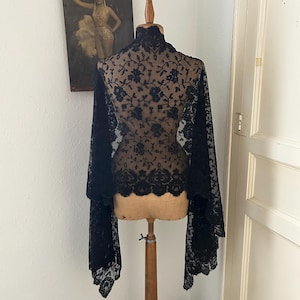 Puede incluir: Un chal de encaje negro con patrón floral drapeado sobre un maniquí de costura beige. El chal presenta un cuello alto, bordados intrincados y un dobladillo festoneado que se extiende por los lados, mostrando una estructura textil delicada.
