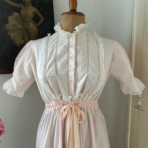 K&ouml;nnte beinhalten: Ein rosa Baumwollkleid mit wei&szlig;er Spitzenborte und einer passenden Schleife. Das Kleid hat einen hohen Kragen und kurze &Auml;rmel.