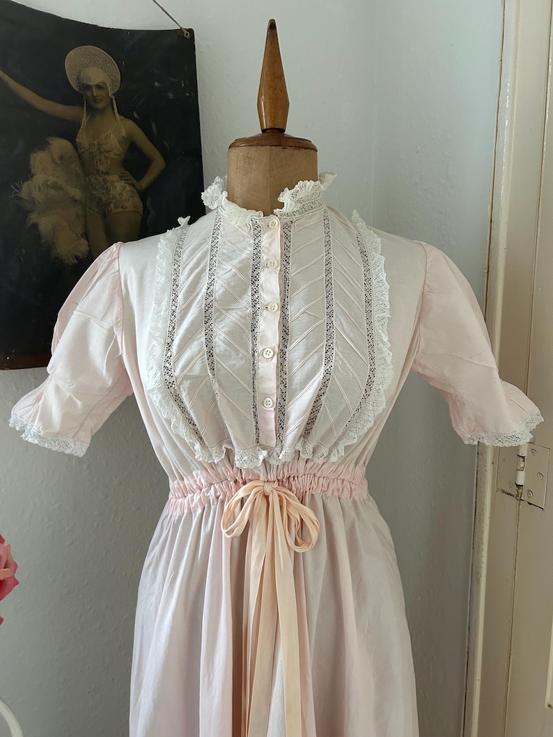 K&ouml;nnte beinhalten: Ein rosa Baumwollkleid mit wei&szlig;er Spitzenborte und einer passenden Schleife. Das Kleid hat einen hohen Kragen und kurze &Auml;rmel.