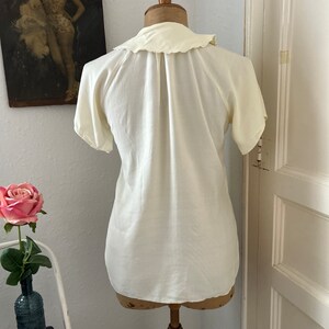 May include: A white, short-sleeved blouse with a ruffled collar. The blouse is on a white mannequin.
