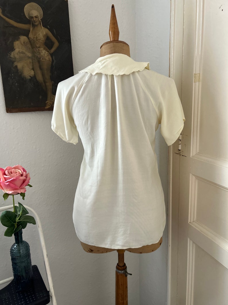 May include: A white, short-sleeved blouse with a ruffled collar. The blouse is on a white mannequin.