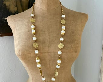 May include: A long gold-tone chain necklace featuring alternating white faux pearl beads and circular gold-tone metal discs with textured surfaces. The necklace is constructed with ornate gold-tone bead caps at the ends of each pearl.