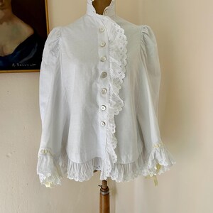 May include: A white blouse with a high ruffled collar and button-down front. The blouse features lace trim along the front, cuffs, and hem. The sleeves are long and slightly puffed at the shoulders. Yellow ribbons accent the cuffs.