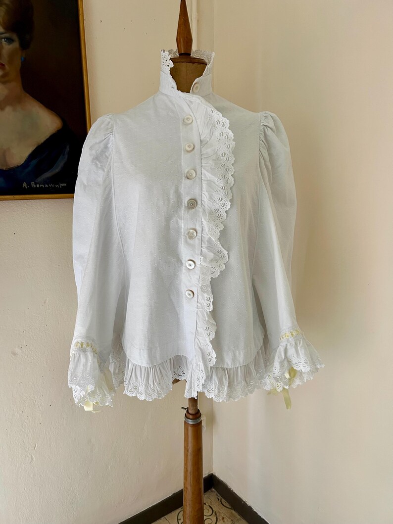 May include: A white blouse with a high ruffled collar and button-down front. The blouse features lace trim along the front, cuffs, and hem. The sleeves are long and slightly puffed at the shoulders. Yellow ribbons accent the cuffs.
