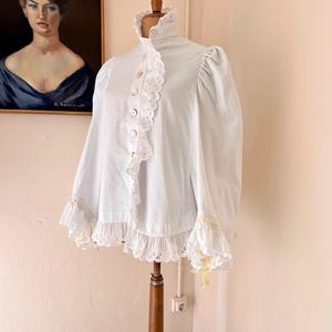 May include: A white vintage blouse with a high ruffled collar and button-down front. The blouse features lace trim along the collar, front, and cuffs, with a touch of yellow ribbon. The sleeves are long and puffy, giving it a Victorian aesthetic.
