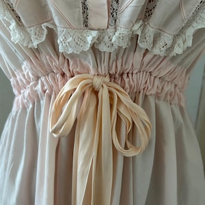 K&ouml;nnte beinhalten: Ein rosa Baumwollkleid mit einer Spitzenborte und einer Satinband-Schleife an der Taille.