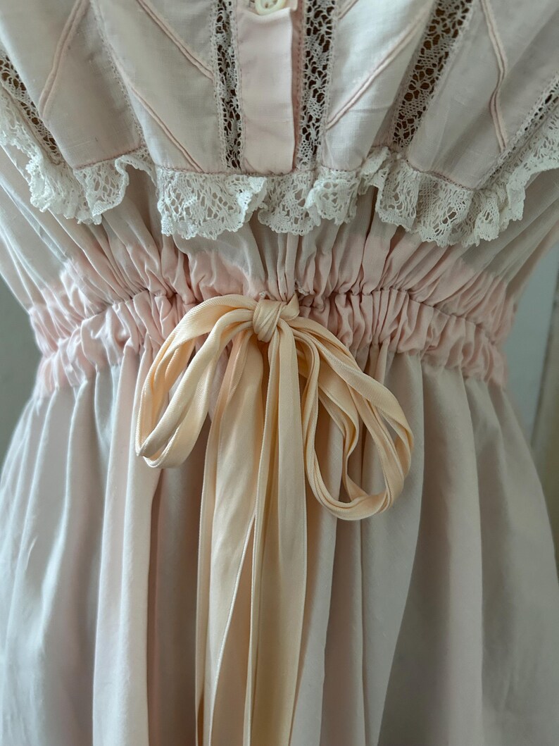 K&ouml;nnte beinhalten: Ein rosa Baumwollkleid mit einer Spitzenborte und einer Satinband-Schleife an der Taille.