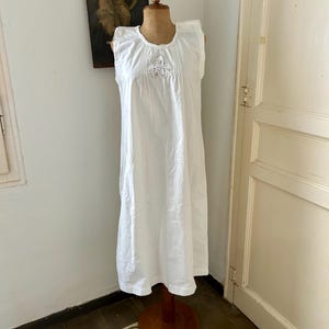 May include: A sleeveless white nightgown featuring a round neckline with vertical pleats and a central panel of intricate floral eyelet embroidery. The garment has a loose, flowing silhouette that extends to the mid-calf, crafted from a lightweight fabric.