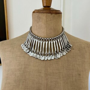 May include: Silver-toned statement necklace with a bib design. The necklace features a chain collar and a series of vertical bars and small circular pendants that hang down. The jewelry is displayed on a mannequin.