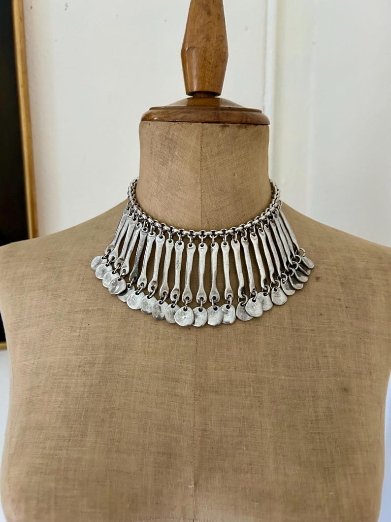 May include: Silver-toned statement necklace with a bib design. The necklace features a chain collar and a series of vertical bars and small circular pendants that hang down. The jewelry is displayed on a mannequin.