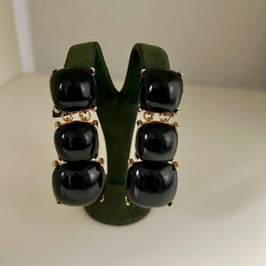 May include: A pair of statement earrings featuring three stacked, glossy black square beads. The beads are connected by gold-toned metal accents. The earrings are displayed on a green velvet earring stand, showcasing their design and color.