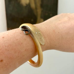 Può includere: Un bracciale rigido in corno liscio, traslucido e color marrone chiaro, caratterizzato da un design a testa di animale intagliata su un'estremità. La testa dell'animale presenta due piccoli inserti circolari bianchi per gli occhi e dettagli incisi.
