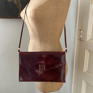 Può includere: Una pochette in pelle di serpente color bordeaux con una chiusura dorata e una lunga tracolla.