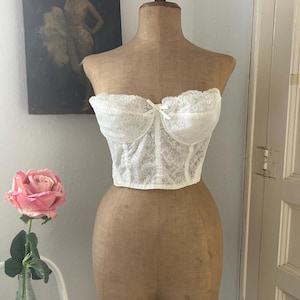 Op de afbeelding: Een wit kanten korset met een sweetheart halslijn en een kleine strik wordt getoond op een vintage paspop. Het korset heeft een delicaat, ingewikkeld kantpatroon. Een roze roos in een glazen vaas staat op de voorgrond.