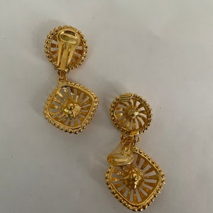 May include: A pair of gold tone clip-on earrings with a square design. The earrings have a delicate, intricate pattern and a beaded edge.