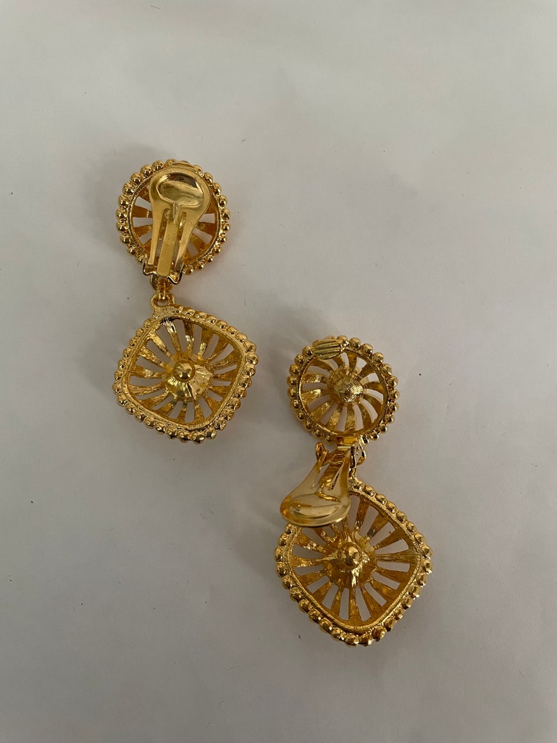 May include: A pair of gold tone clip-on earrings with a square design. The earrings have a delicate, intricate pattern and a beaded edge.