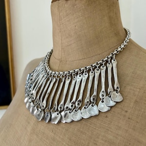May include: Silver-toned statement necklace with a chain collar and a bib of vertical bars and small, heart-shaped pendants. The necklace is displayed on a tan dress form, showcasing its design and texture.