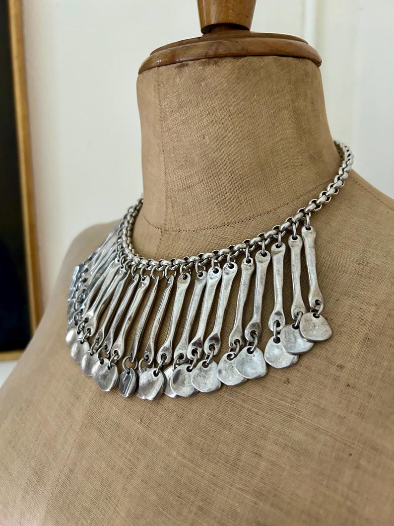 May include: Silver-toned statement necklace with a chain collar and a bib of vertical bars and small, heart-shaped pendants. The necklace is displayed on a tan dress form, showcasing its design and texture.