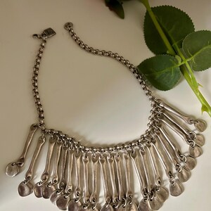 May include: Silver-toned necklace with a chain and pendant design. The necklace features a series of elongated, spoon-shaped pendants with rounded bottoms. The necklace is displayed on a white surface with a red rose in the background.