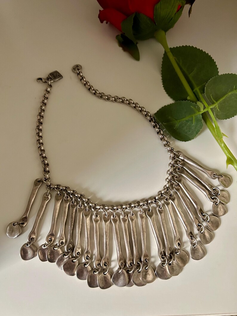 May include: Silver-toned necklace with a chain and pendant design. The necklace features a series of elongated, spoon-shaped pendants with rounded bottoms. The necklace is displayed on a white surface with a red rose in the background.