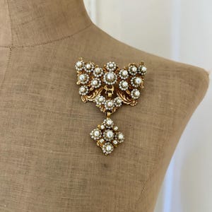 May include: A vintage gold-tone brooch with faux pearls. The brooch has an ornate design with a central cluster of pearls and a dangling pendant. The brooch is displayed on a mannequin.