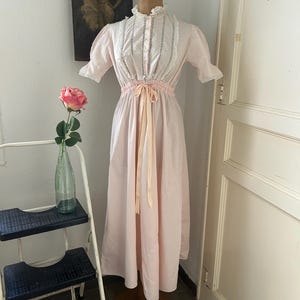 K&ouml;nnte beinhalten: Ein langes, rosafarbenes, vintage Nachthemd mit wei&szlig;en Spitzenbesatz und einer Satinband-Schleife an der Taille.