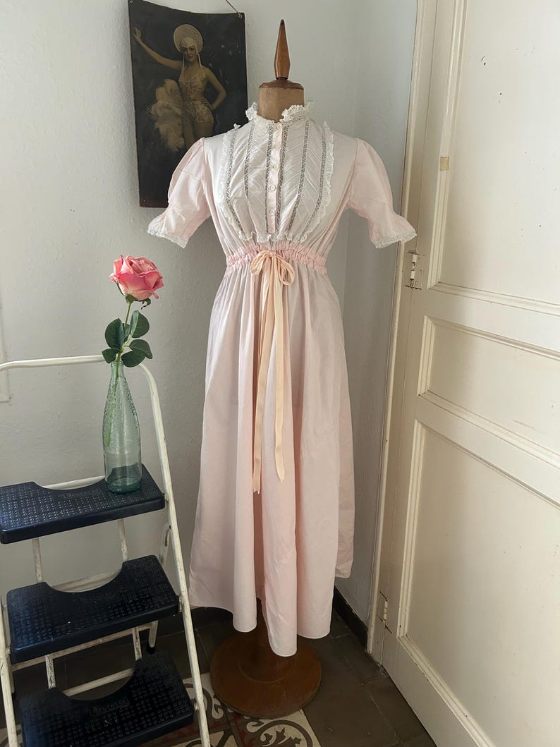 K&ouml;nnte beinhalten: Ein langes, rosafarbenes, vintage Nachthemd mit wei&szlig;en Spitzenbesatz und einer Satinband-Schleife an der Taille.