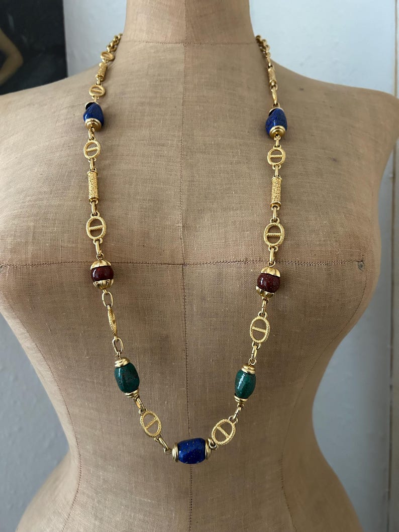 May include: A gold chain necklace with blue, green, and red oval-shaped beads. The chain features a repeating pattern of interlocking circles.