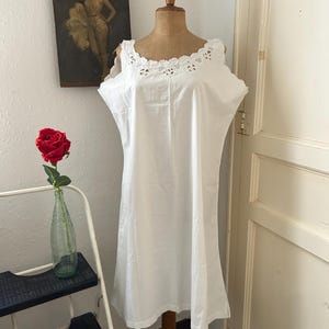 Könnte beinhalten: Ein weißes Vintage-Unterkleid mit zarter Spitzenborte am Ausschnitt und an den Schultern. Das Kleid ist auf einer Holzpuppe ausgestellt. Eine rote Rose in einer grünen Glasflasche und ein Vintage-Poster befinden sich im Hintergrund.