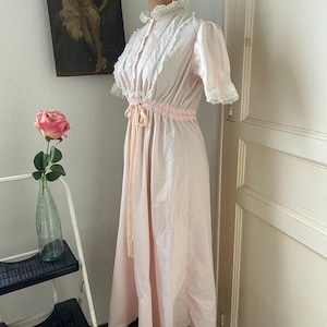 K&ouml;nnte beinhalten: Ein langes, rosa, baumwollenes Nachthemd mit einer wei&szlig;en Spitzenborte am Ausschnitt und an den &Auml;rmeln. Das Nachthemd hat eine Kordeltaille und eine Schleife.