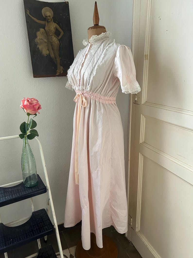 K&ouml;nnte beinhalten: Ein langes, rosa, baumwollenes Nachthemd mit einer wei&szlig;en Spitzenborte am Ausschnitt und an den &Auml;rmeln. Das Nachthemd hat eine Kordeltaille und eine Schleife.