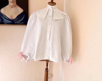 May include: A white blouse with a high collar and button-down front is displayed on a wooden mannequin. The sleeves are gathered at the cuffs, which are adorned with pink ribbons. A framed portrait hangs in the background.