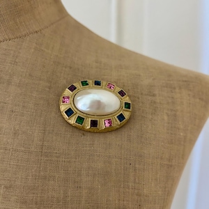 May include: Gold-toned oval brooch with a central pearl and a border of colorful square gemstones. The brooch is displayed on a tan fabric surface, showcasing its vintage design and vibrant colors.