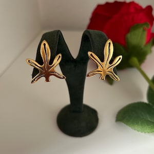 Puede incluir: Un par de pendientes de hoja de oro en un expositor negro. Los pendientes son simples y elegantes.