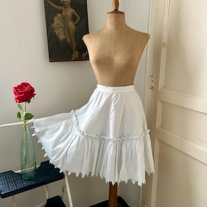Op de afbeelding: Een witte, geplooide rok tentoongesteld op een vintage paspop. De rok heeft een geschulpte zoom en een gerimpelde taille. Een vintage poster hangt aan de muur. Een rode roos in een groene glazen fles staat op een kleine zwarte kruk.