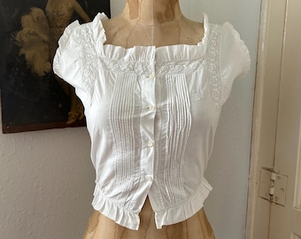 May include: A white short-sleeved blouse featuring a square neckline, delicate lace trim along the shoulders, vertical pleats down the front, and a button-down closure. The garment is finished with ruffled edges at the sleeves and hemline.
