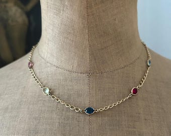 May include: A gold-toned chain necklace featuring evenly spaced, bezel-set gemstones in various colors including deep blue, bright red, light green, and pink. The delicate metal links connect each individual stone, creating a simple, elegant piece of jewelry.