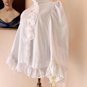 May include: A white vintage blouse with a high ruffled collar and lace trim. The blouse features button closures down the front and ruffled sleeves. A yellow ribbon is tied on the sleeve. The blouse is displayed on a wooden mannequin.