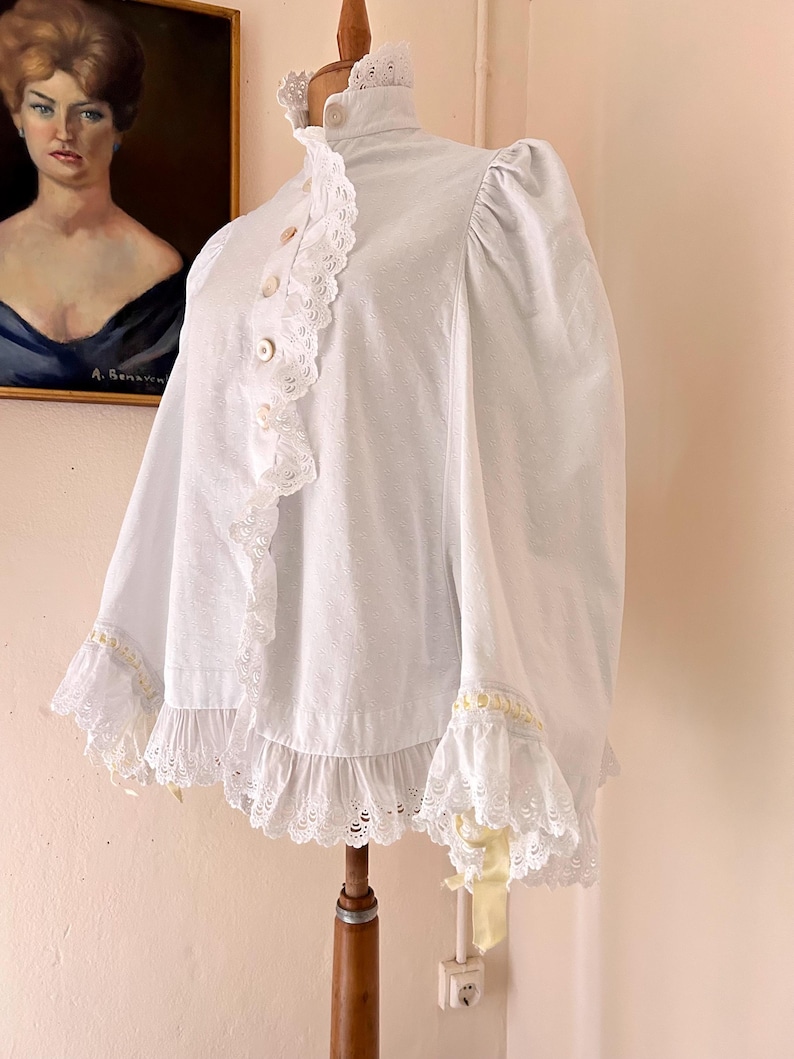 May include: A white vintage blouse with a high ruffled collar and lace trim. The blouse features button closures down the front and ruffled sleeves. A yellow ribbon is tied on the sleeve. The blouse is displayed on a wooden mannequin.