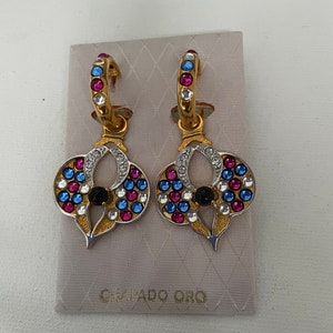 Pode incluir: Um par de brincos de argola dourados com um pingente em forma de coração. O pingente é decorado com pedras de cristal coloridas e uma pedra preta no centro. Os brincos estão embalados em uma bolsa branca com o texto "Camaldo Oro" impresso nela.