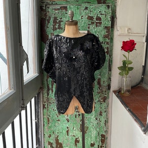 Puede incluir: Un top negro de lentejuelas con cuello redondo y mangas cortas. El top está en un maniquí sobre un fondo verde y blanco.