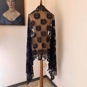 Può includere: Uno scialle di pizzo nero con un motivo floreale è drappeggiato su un manichino di legno. Lo scialle ha un bordo smerlato ed è esposto davanti a un dipinto di una donna.