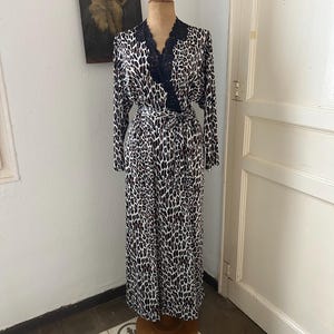 Op de afbeelding: Een kamerjas met lange mouwen en een vloerlange lengte, voorzien van een luipaardprint in bruin, zwart en wit. Het kledingstuk heeft een zwarte kanten rand langs de V-hals en een bijpassende stoffen ceintuur in de taille, getoond op een houten paspop.