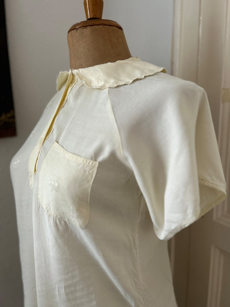 May include: A white, short-sleeved blouse with a Peter Pan collar and a single pocket on the left side.