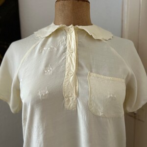 May include: A white cotton shirt with a scalloped collar and short sleeves. The shirt has a button-up front and a small pocket on the left chest. The shirt is embroidered with small, white flowers.