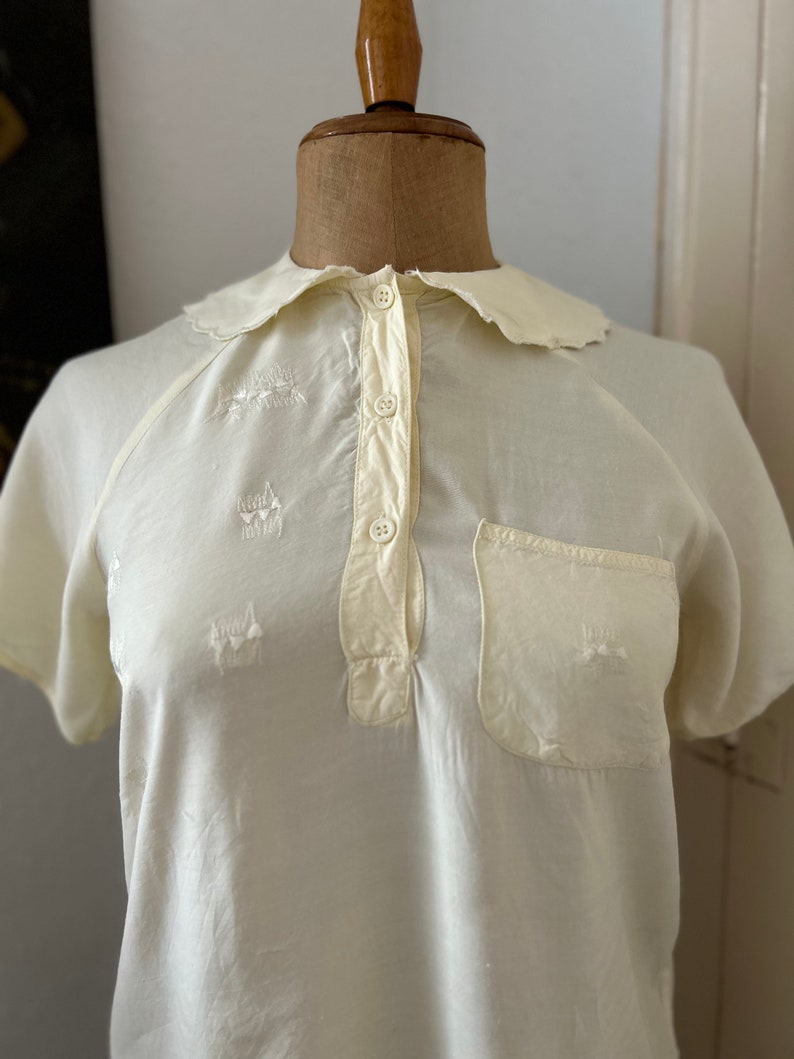 May include: A white cotton shirt with a scalloped collar and short sleeves. The shirt has a button-up front and a small pocket on the left chest. The shirt is embroidered with small, white flowers.