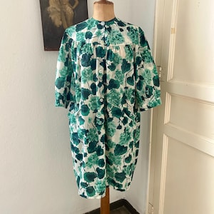 Könnte beinhalten: Ein knielanger Hausmantel mit weißem Grund und einem leuchtend grünen Blumenmuster. Das Kleidungsstück hat einen Rundhalsausschnitt, kurze Ärmel und eine durchgehende Knopfleiste mit hellen Knöpfen. Es verfügt über zwei seitliche Eingrifftaschen.