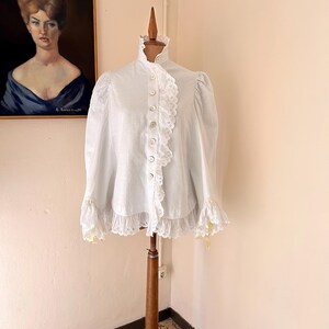 May include: A white blouse with a high ruffled collar and lace trim. The long-sleeved top features a button-down front and is displayed on a wooden dress form. The blouse has lace detailing on the cuffs and down the front.