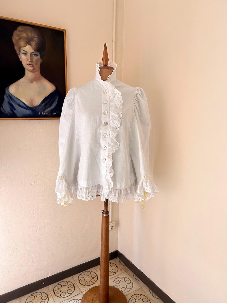 May include: A white blouse with a high ruffled collar and lace trim. The long-sleeved top features a button-down front and is displayed on a wooden dress form. The blouse has lace detailing on the cuffs and down the front.