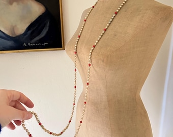 May include: A long, layered necklace with faux pearls and red beads. The necklace is draped on a vintage dress form. The pearls are off-white, and the red beads are evenly spaced throughout the strand. A painting hangs in the background.